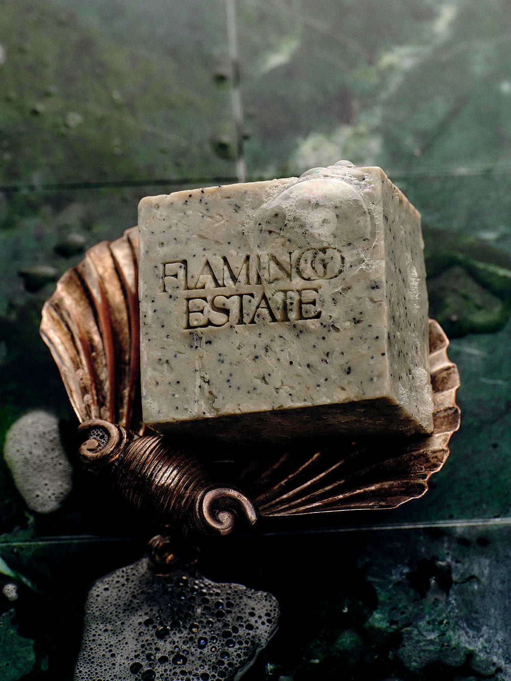 Bar of soap with 'Flamingo Estate' branding on a decorative holder against a green leafy background.