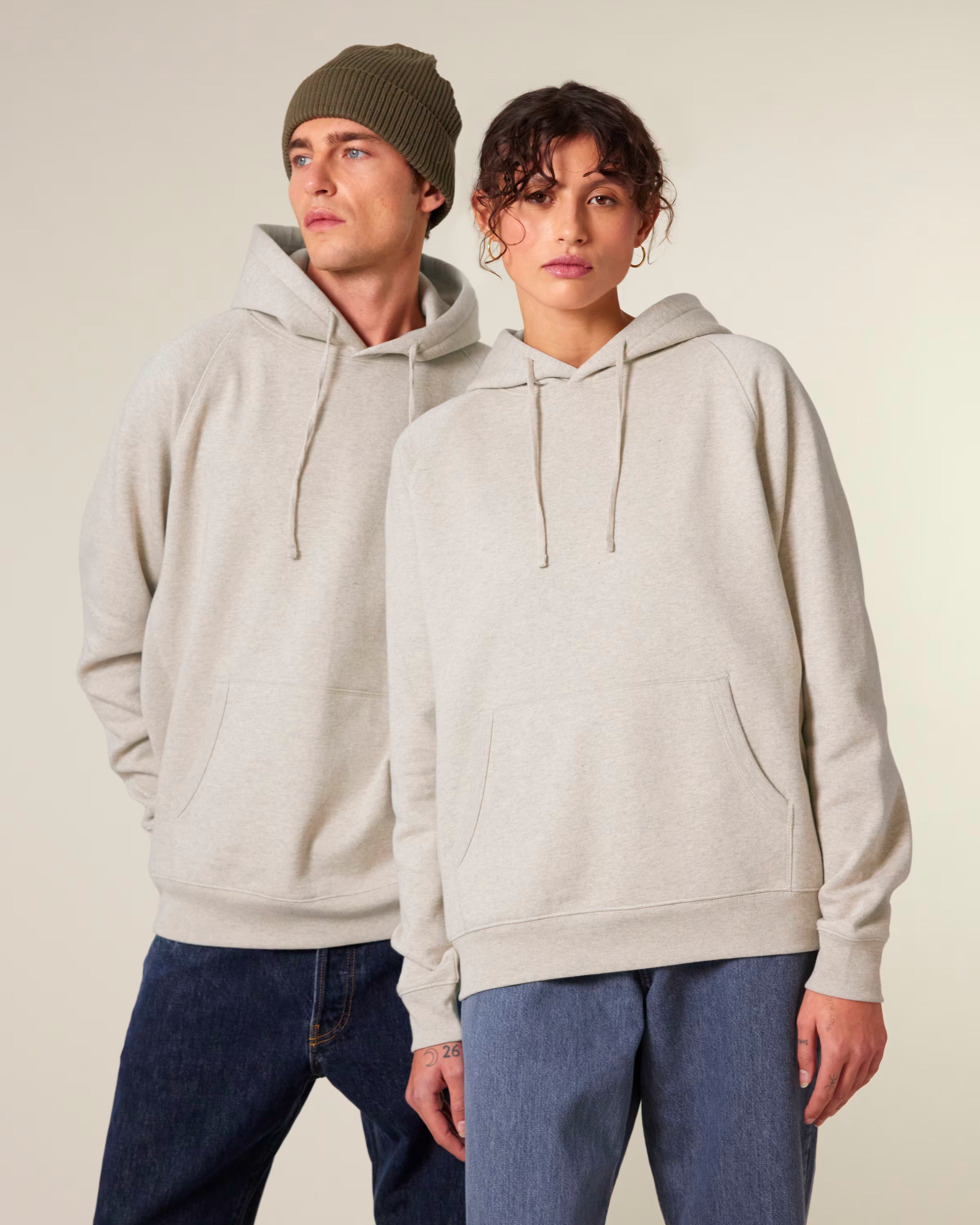 Two people wearing beige unisex cotton hoodies and blue jeans against a plain background