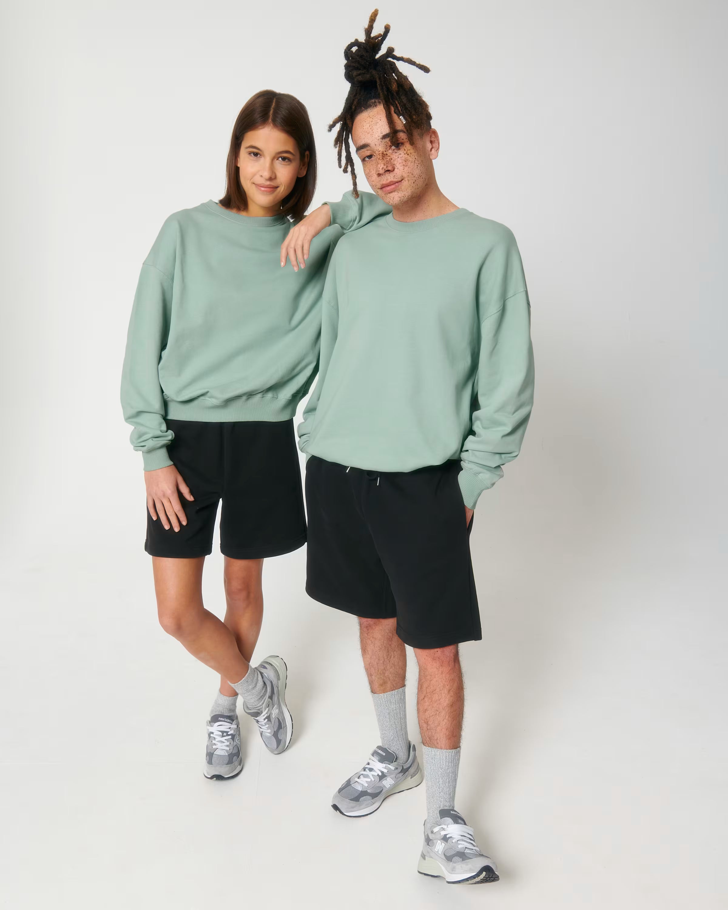 Two people wearing matching green unisex cotton sweatshirts and black shorts on a white background
