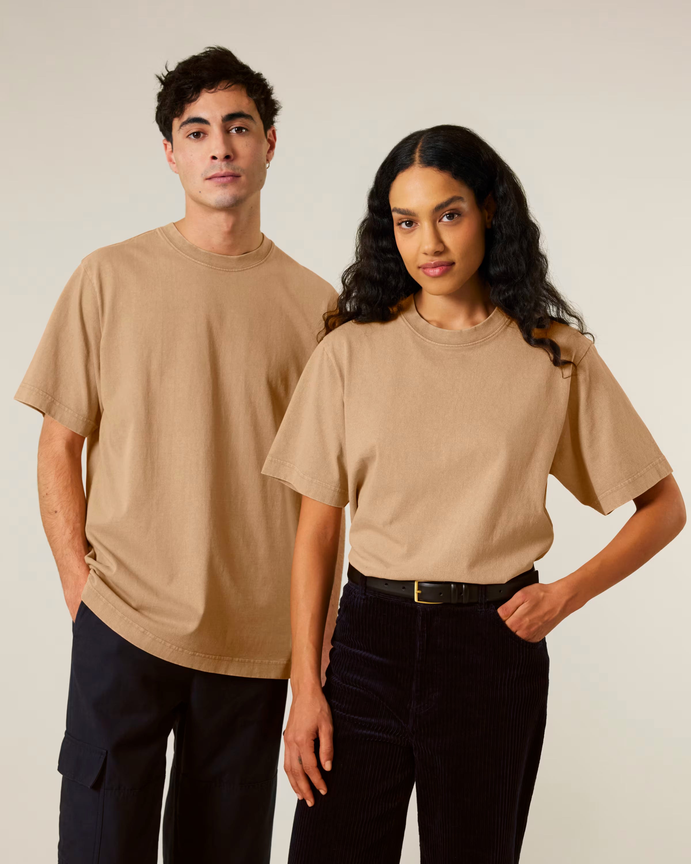 Two people wearing beige unisex cotton t-shirts and black pants against a plain background