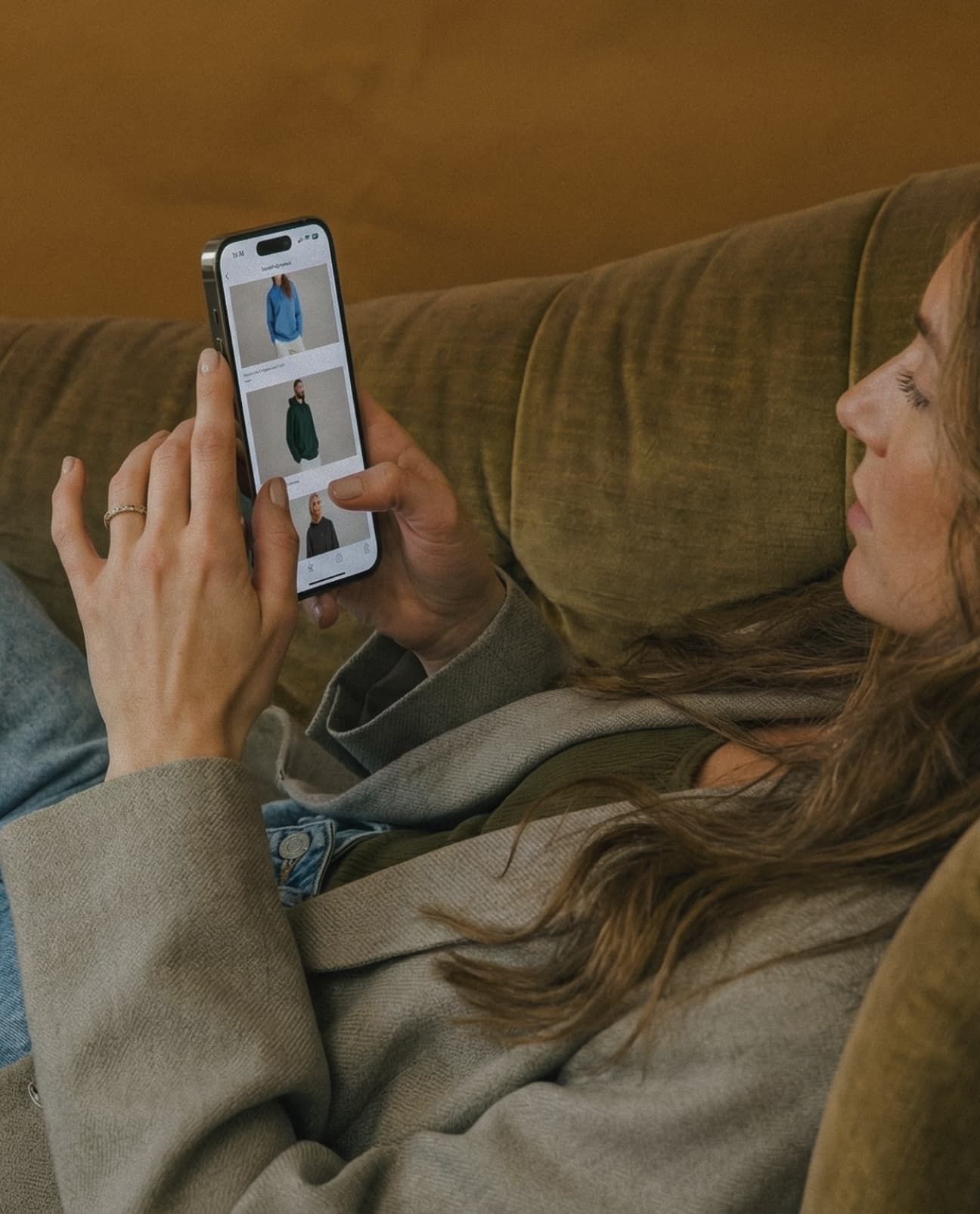 Woman sitting on a couch looking at a phone with clothing images displayed.