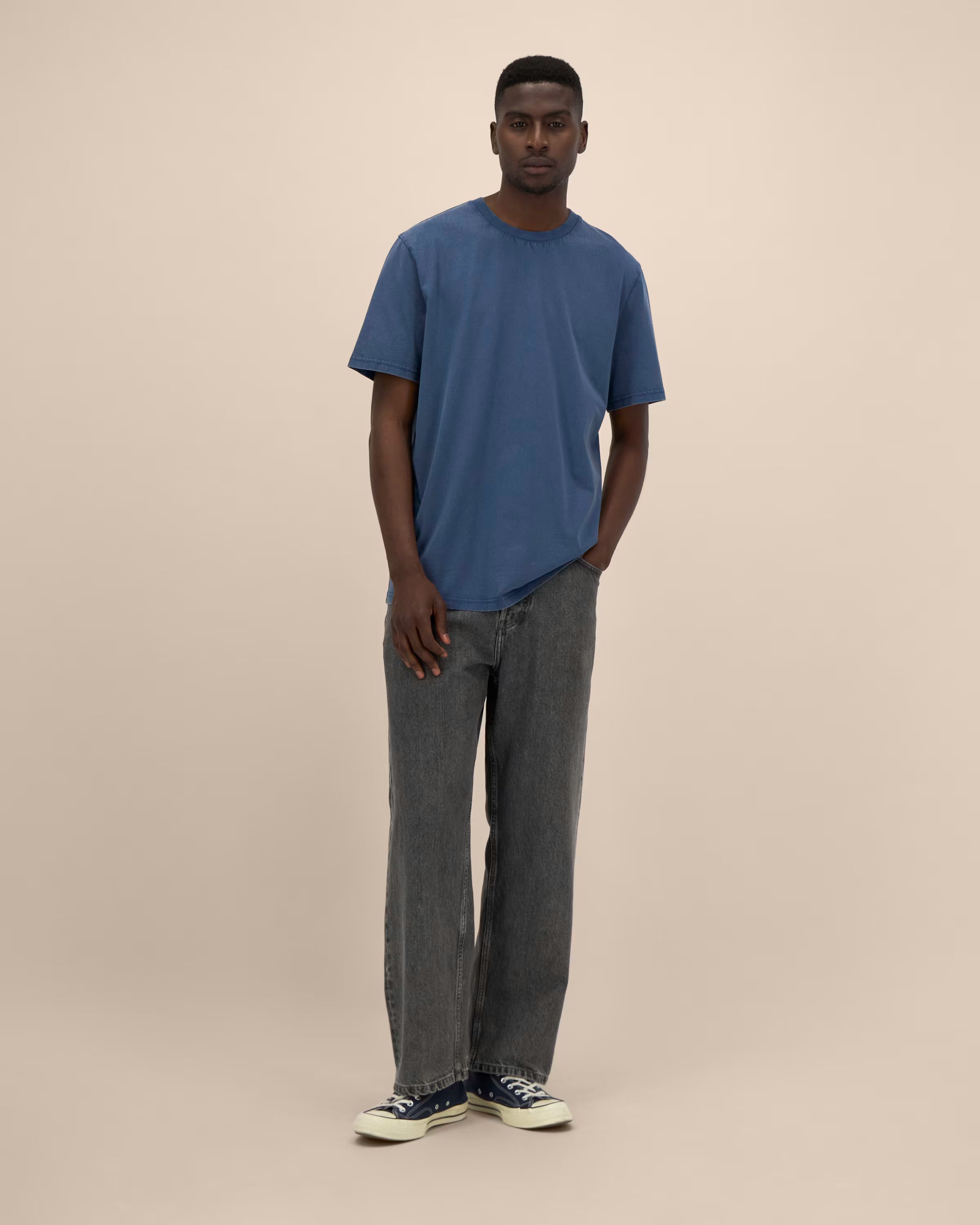 Man wearing a blue unisex cotton t-shirt and gray jeans standing against a beige background