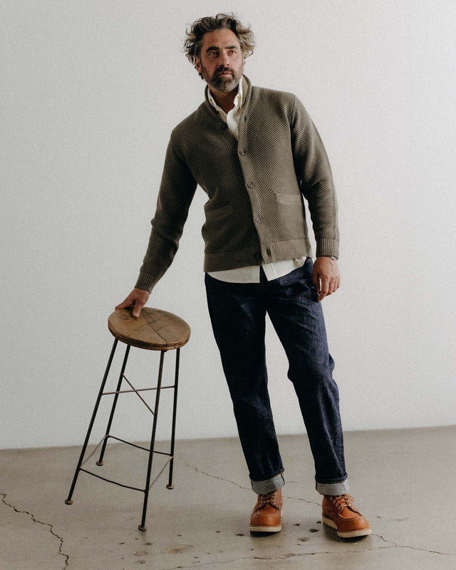 Man wearing a green cardigan, white shirt, and dark jeans standing next to a wooden stool against a plain wall.